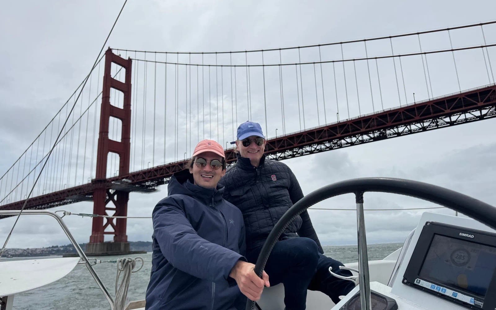 Two guests at the wheel of a sailboat directly beneath the Golden Gate Bridge on an overcast day